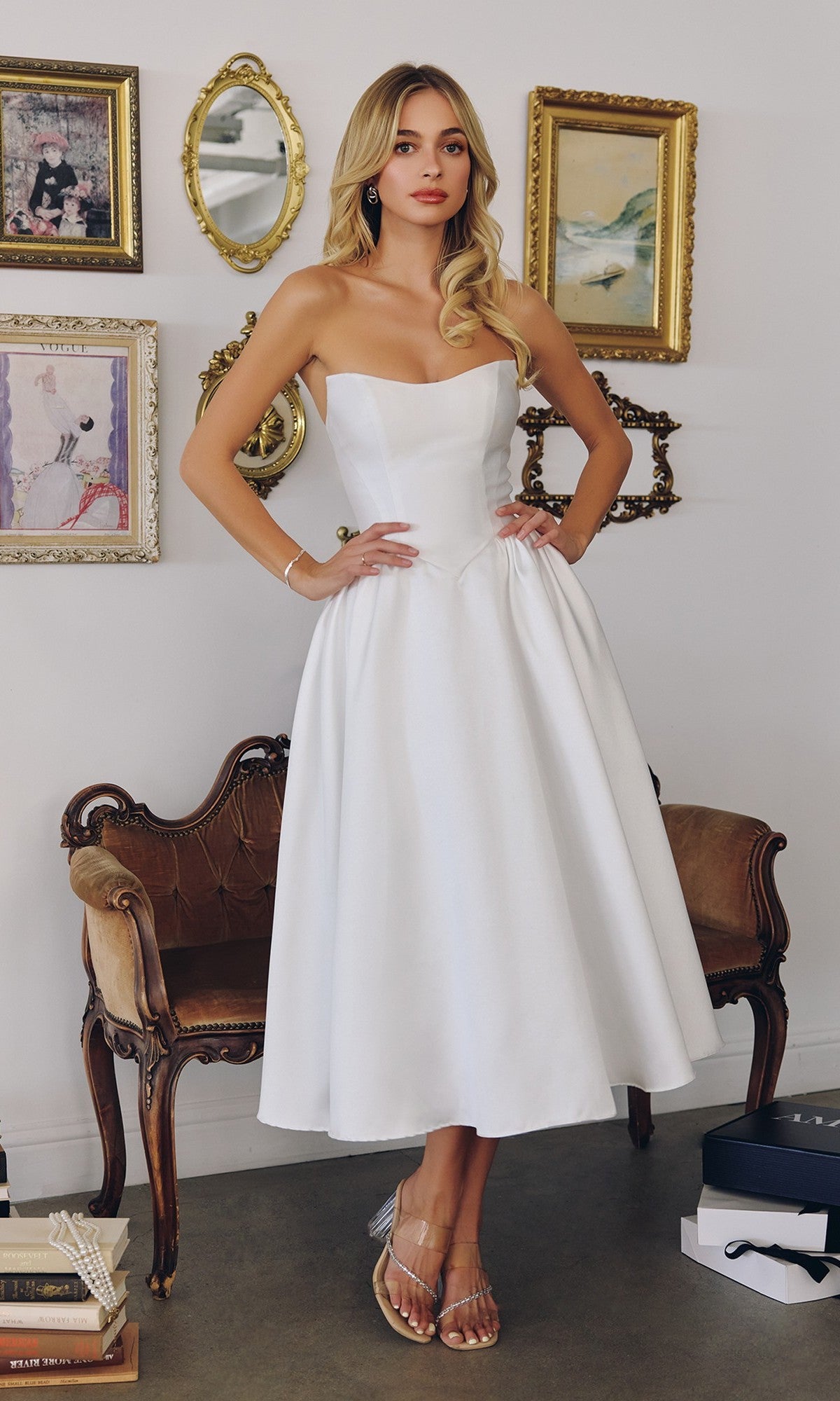 A woman in the Off-White Midi Prom Dress: Amoris T822W stands indoors near a vintage chair, with framed art on the wall behind her. She wears clear sandals; the dress features a lace-up back.